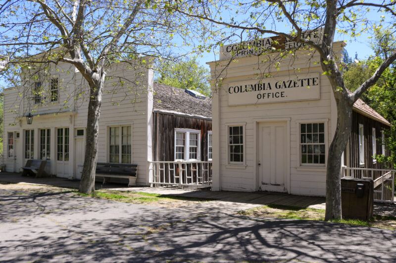 two historic white clapboard buildings on a street with several trees in front, the closest building says Columbia Gazette on the sign, under a clear blue sky at Columbia State Historic Park