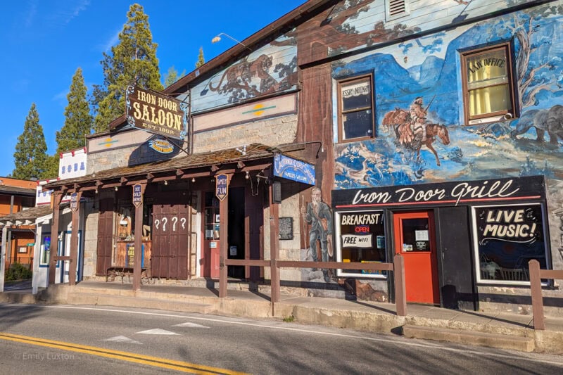 exterior of the Iron Door Saloon in Groveland, a large buildign with a mural showing a cowboy on a horse on a blue background. There is an empty street in front and blue sky above. Groveland high street in Tuolumne County California