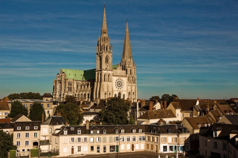 The Our Lady of Chartres cathedral, a large grey stone building with two towers and a circular cnetral window between the two. It is on a hill surrounded by houses in the centre of Chartres against a clear blue sky. 