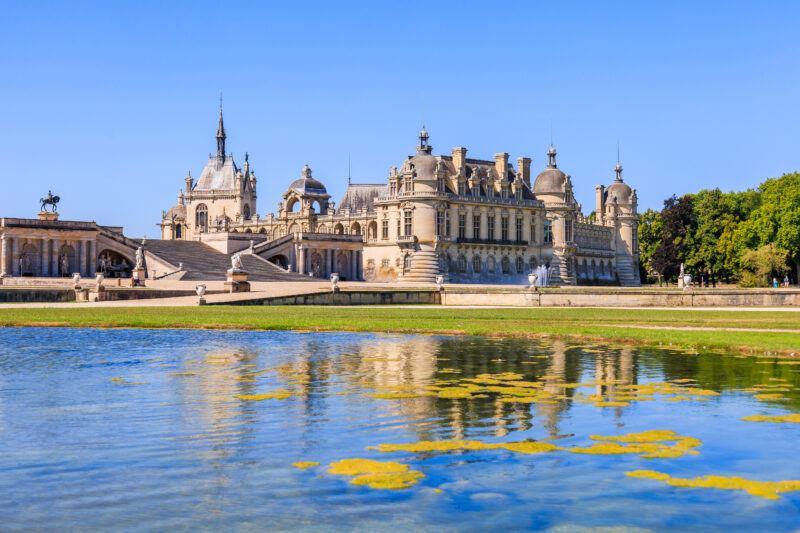 View of the Chateau de Chantilly from the garden with a large lage in the foreground and the chateau reflected in the lake. It is a sunny day with clear blue sky overhead. 