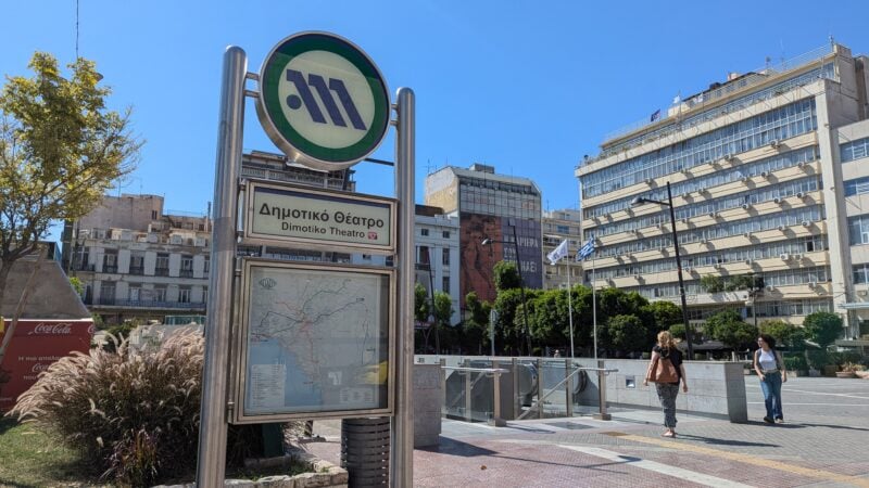 sign for Dimotiko Theatro metro station. At the top of the sign there is a white circle containing the blue metro logo, and behind are steps descending into the station. In a square with a large tower block in the background and clear blue sky overhead. 