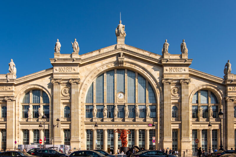 Exterior of Gare du Nord station in Paris, France, a large grey stone building with three large arched windows on the front and several pillars, against a clear blue sky. Best day trips from Paris. 