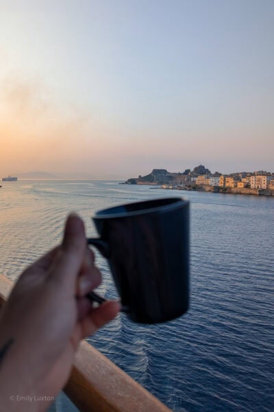 hand holding a coffee cup over a balcony railing with a view of the sea behind and a low headland out of focus behind that, just after sunrise with a pink haze in the sky and golden glow on the white buildings on the headland. 