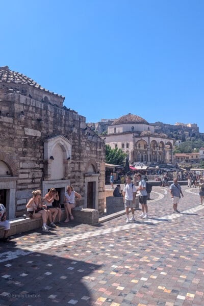 old stone church on the edge of a large square with pink stone tiles, there is an ottoman church in the background with white walls and an orange tiled domed roof, and a grassy hill behind that with clear blue sky overhead. 