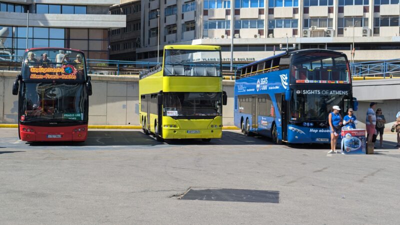 three double decked buses in a car park with tower blocks behind, from left to right they are red, yellow and blue. Hop on hop off bus tour from Piraeus Port in Athens. 