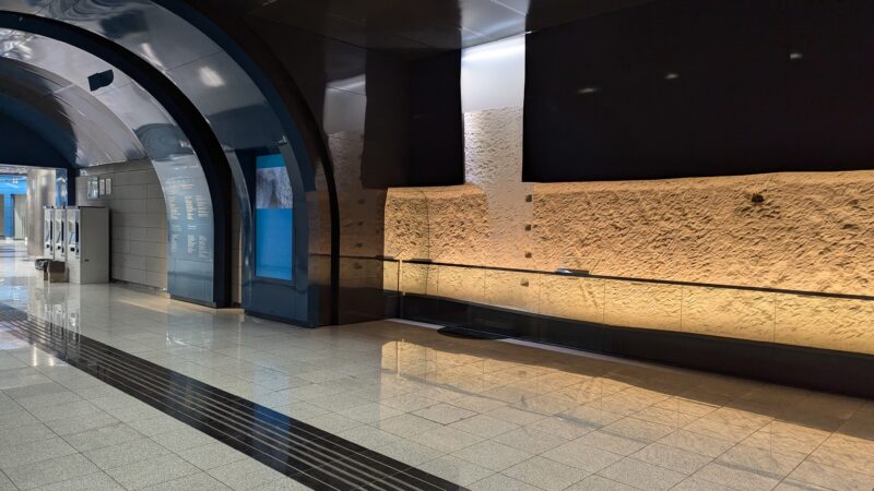 Ancient stone wall with holes lit up by orange lights behind glass inside Dimotiko Theatro metro station in Athens, with an arched ceiling overhead and white marble tiles on the floor. 