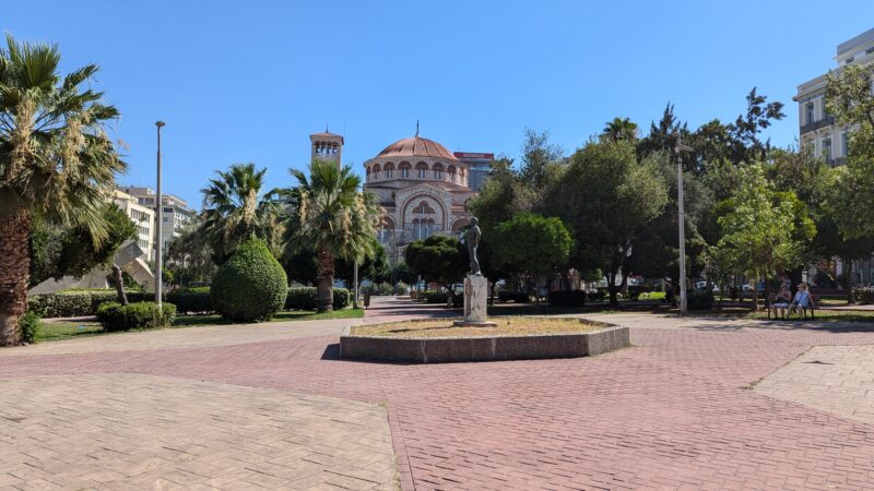 statue of a man on a grey stone plinth at the centre of a large square with pink stone tiles in a park surrounded by green leafy trees, with a large church in the background with a red tiled domed roof