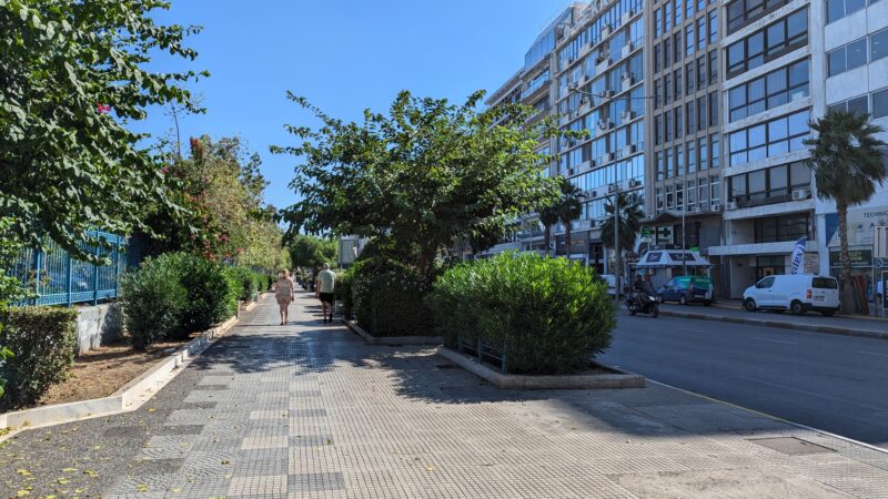 wide grey pavement or sidewalk with a row of green leafy trees planted between it and the road and a row of tower blocks on the other side of the road