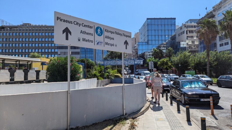 pavement next to a road with a large recangular sign reading Piraeus City Centre, Athens, Metro and an arrow pointing forward