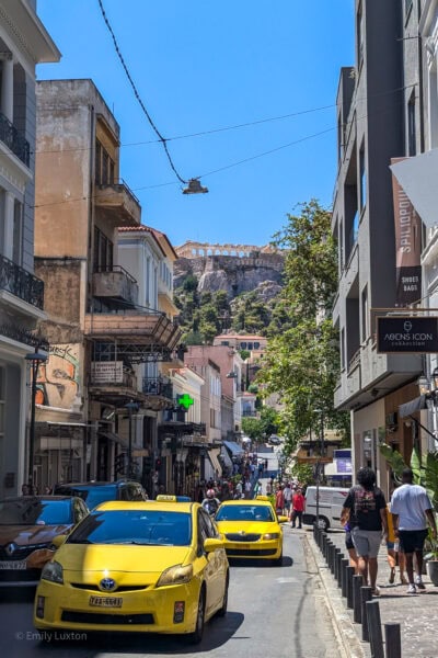 two yellow taxis in a narrow road between tall buildings in Athens city centre with a view of the ancient Acropolis on a hilltop behind and clear blue sky overhead