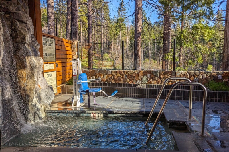 large rectangular hot tub set into the ground surroudnedby grey tiles with a rock wall ont he left where a mini waterfall runs into the hot tub. Beyond the tub is a view of pine forest on a sunny day with clear blue sky behind the trees. 