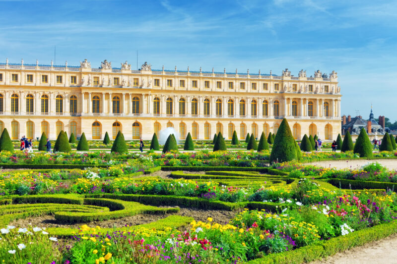 Pond in front of the Royal residence at Versailles near Paris in France, surrounded by green lawnsa dn decorative hedges, with the yellow exterior of the palace behind and clear blue sky above
