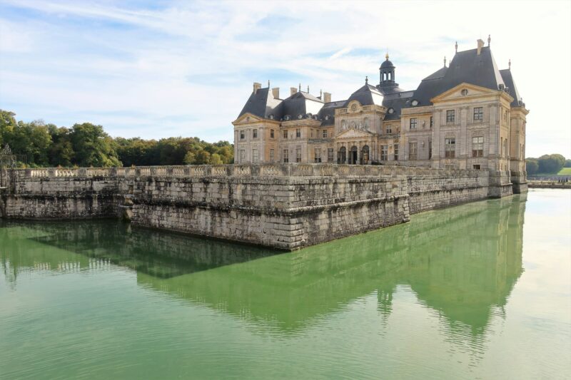green lake in front of a large grey stone chateau with a dark grey roof on a sunny day with a cloudy sky above. 