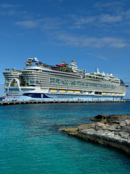 very large white and blue cruise ship at sea with turquoise water and rocks in foreground and blue sky above