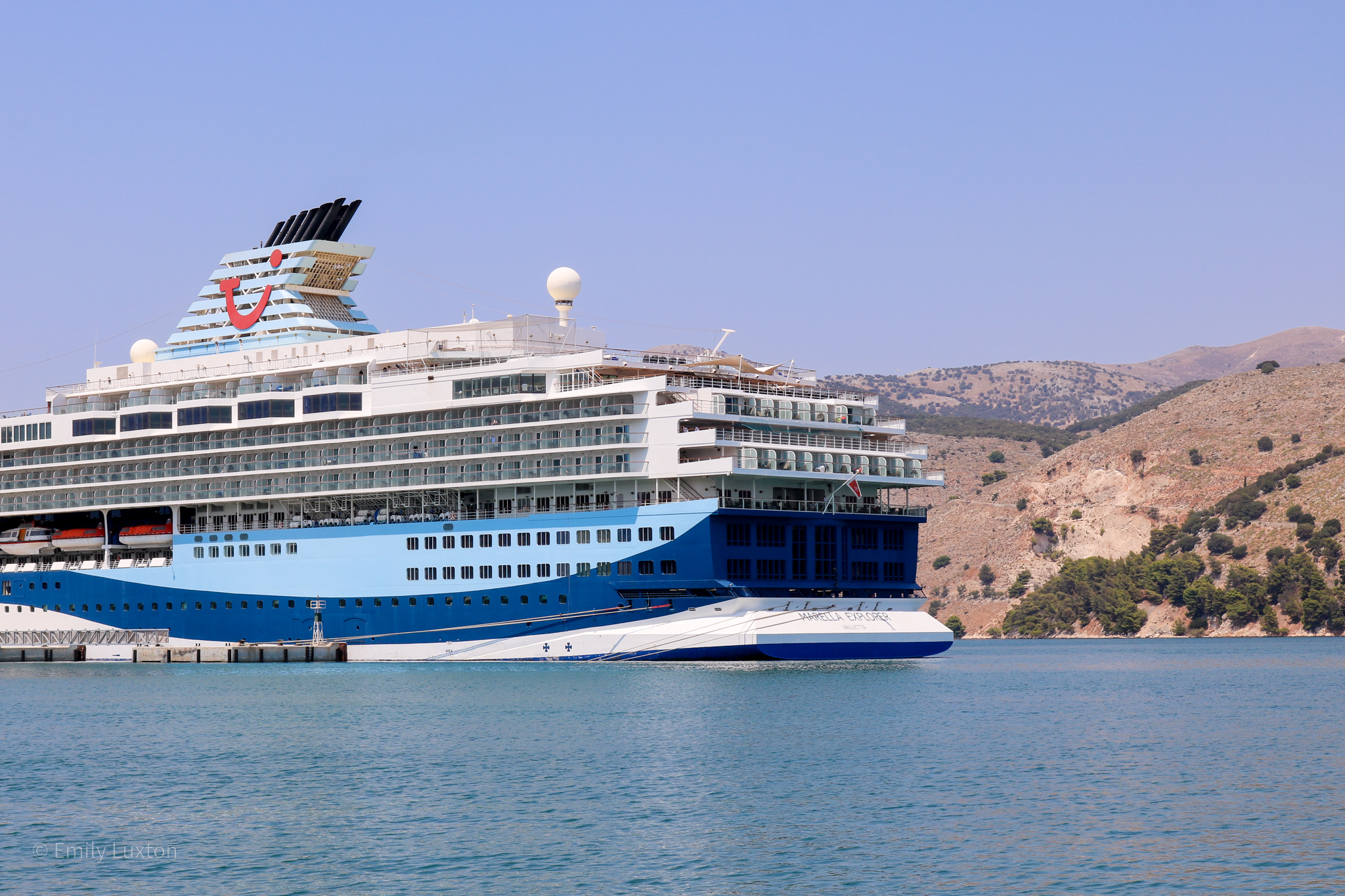 Marella Explorer cruise ship in a bay in Kefalonia with a yellow-green grassy hill behind. The ship has a two toned blue base, white exterior and a large cyan funnel with the Tui logo on it - a red smile with a dot above one end.