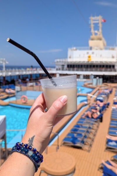 hand holding a plastic tumbler of pina colada with a black paper straw in front of the pool deck of a cruise ship with the ship and pools out of focus in the background. There is a purple watch strap and a small black tatoo of a pterodactyl on the wrist.