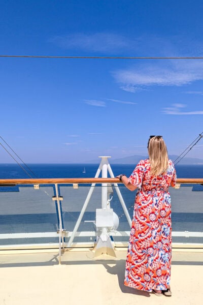 emily wearing a long orange dress with blue and white swirls standing on the deck of a cruise ship looking away from the camera over the balcony towards blue sea. It is a very sunny day with clear blue sky above.