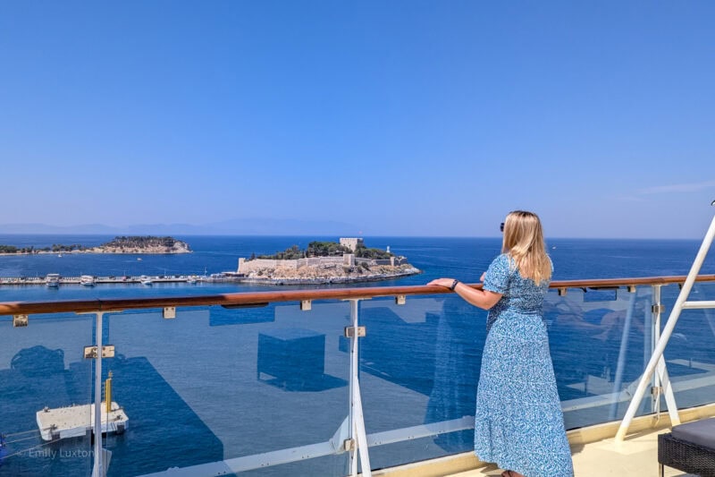 emily standing on the deck of a cruise ship looking away from the camera over the balcony towards blue sea and a small island with a castle on it. it is a very sunny day with clear blue sky above.