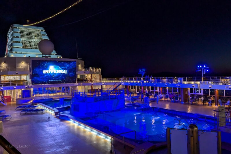 top deck of a cruise ship at night with a pool lit up in blue and a big screen overlooking the pool. The Universal logo is on the screen. Marella Explorer cruise ship review. 