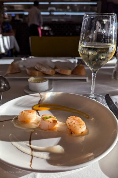 white plate with three scallops surrounded by a smudge of cream coloured sauce, there is a glass of white wine on the table behind and white tablecloth on the table.