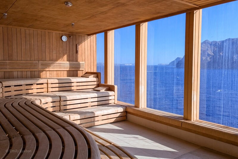 large empty sauna with curved wooden benches in front of large windows with a view of the ocean and a rocky island in the distance - Marella Explorer Ocean Spa.