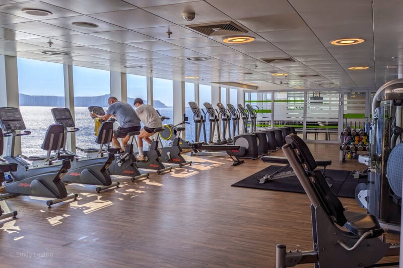 gym with laminated floor and white tiled cieling with a row of exercise bikes and running machines lined up in front of a row of large windows wiht a view of the sea and a rocky island in the distance.