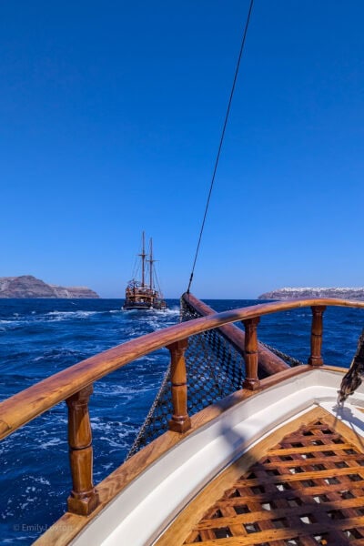 prow of a wooden ship sailing in very bridght blue sea wtih another wooden tall ship in the distance in front and two low rocky island beyond that. It is a very sunny day with clear blu sky above.