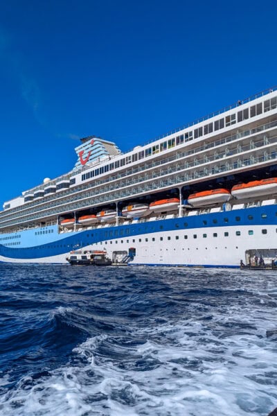 Close up of the side of the Marella Explorer cruise ship with blue sea in front. The ship has a two toned blue base, white exterior and a large cyan funnel with the Tui logo on it - a red smile with a dot above one end.