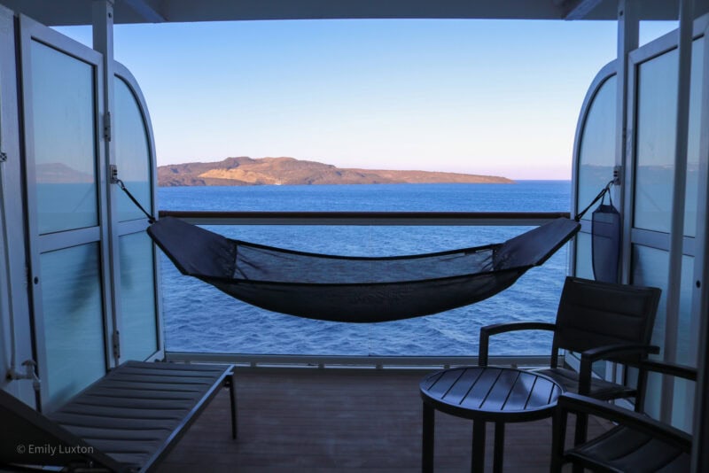 balcony on the side of a cruise ship with a small circular table and a chair next to a hammock strung up between the two walls, the furniture is all silhouetted against the blue sea behind the balcony's glass wall, and beyond the sea is a small brown rocky island.