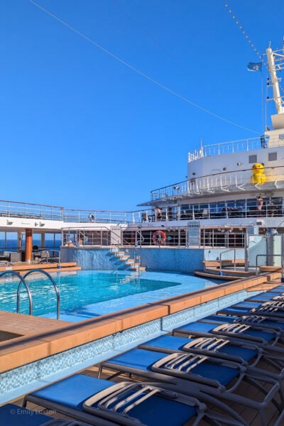 small rectangular pool with very bright cyan tiles on the deck of a cruise ship surrounded by blue sun loungers on a very sunny day with clear blue sky overhead.