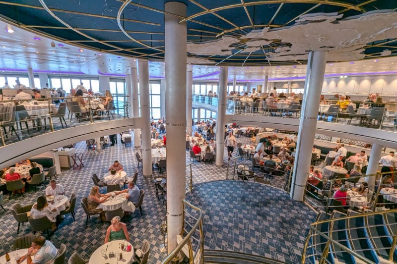 Interior of the main dining room on the Marella Explorer cruise ship, looking out from the central staircase across the dining room which is split over two levels and has a circular blue ceiling with a map of the worlf on it, the lower floor has blue pattended carpet and both levels are full of circular tables with groups of people dining at them.