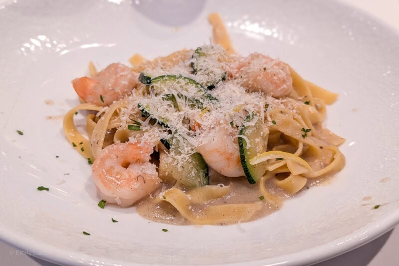 close up of a white plate with spaghetti, courgette slices and prawns in a creamy sauce covered with parmesan shavings.