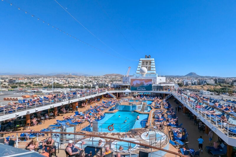 Looking out over the pool deck of the Marella Explorer cruise ship with two small rectangular swimming pools in the centre surrounded by blue sun loungers. There are a large number of people on the deck and it looks very busy and crowded. There is a view of Athens behind the ship and clear blue sky overhead.