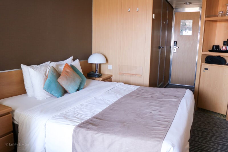 Interior of a cruise ship cabin with one brown wall and one wall clad in light coloured wood. There is a double bed with white sheets, a grey throw and three small cyan cusions in front of the two white pillows. Behind the bed a small cupboard and some shelves are built into the wall.