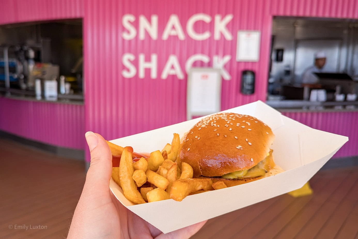 hand holding a white paper box with a burger and chips in it in front of a pink corrugated iron wall with the words Snack Shack printed on it in white.