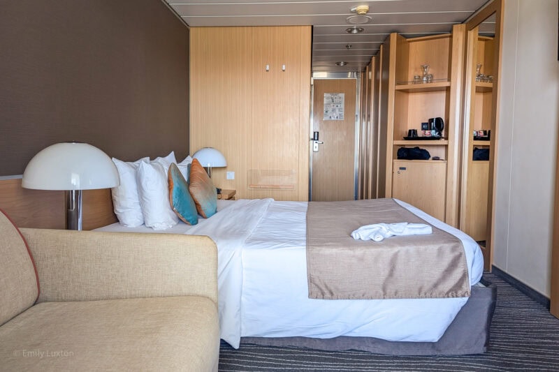 Interior of a cruise ship cabin with one brown wall and one wall clad in light coloured wood. There is a double bed with white sheets, a grey throw and three small cyan cusions in front of the two white pillows. Behind the bed a small cupboard and some shelves are built into the wall. Marella Explorer balcony cabin review. 