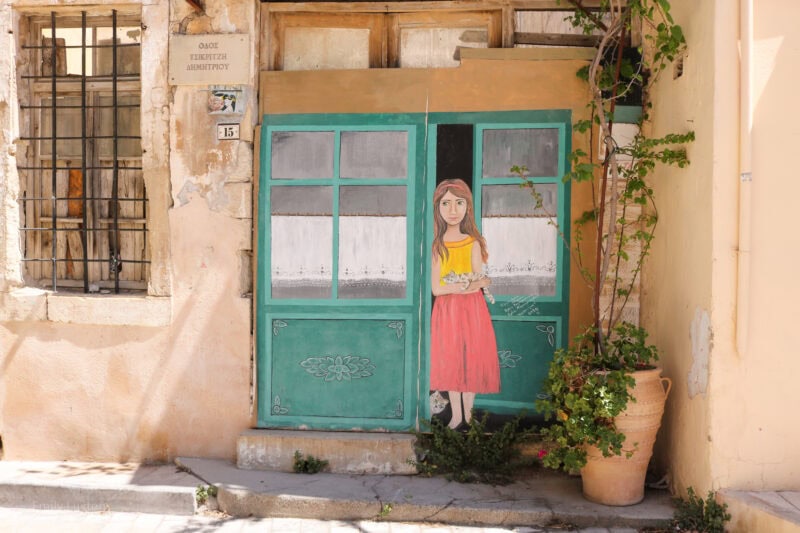 yellow wall with a large painted mural depicting green doors with a painted young girl standing in the open doorway, she is wearing a pink skirt and yellow top and has long brown hair and fair skin.