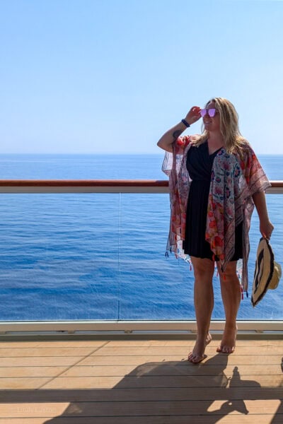 emily standing on a cruise ship balcony with wooden floor and a glass wall with the clear blue sea behind. Emily is wearing a black wrap mini dress and a long orange patterned shawl and is holding a floppy beige sunhat in her left hand and holding onto the side of her sunglasses with the other.