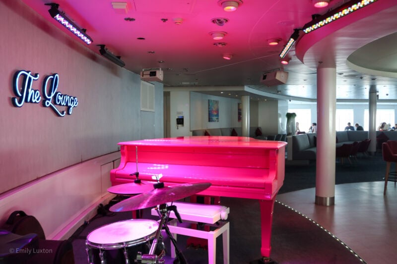 Bright pink piano on a stage with a bar out of focus behind it, the neon sign on the wall says The Lounge.