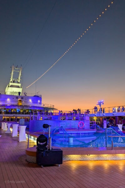 Top deck of a cruise ship at night not long after sunset with an orange glow at the bottom of the sky. The deck is lit by orange lighting and there is a small rectangular pool lit up in blue.