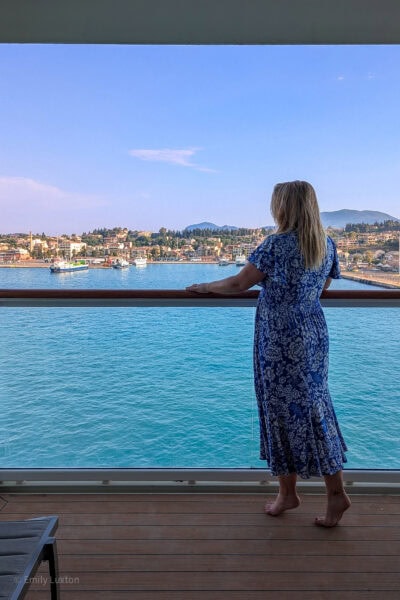 emily wearing a long blue dress with white swirls standing on the balcony of a cruise ship looking away from the camera over the balcony towards blue sea with a view of corfu port beyong that. It is a very sunny day with clear blue sky above.