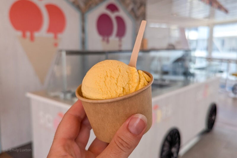 hand holding a brown cardboard tub of yellow ice cream with a white gelato stand out of focus behind it. 