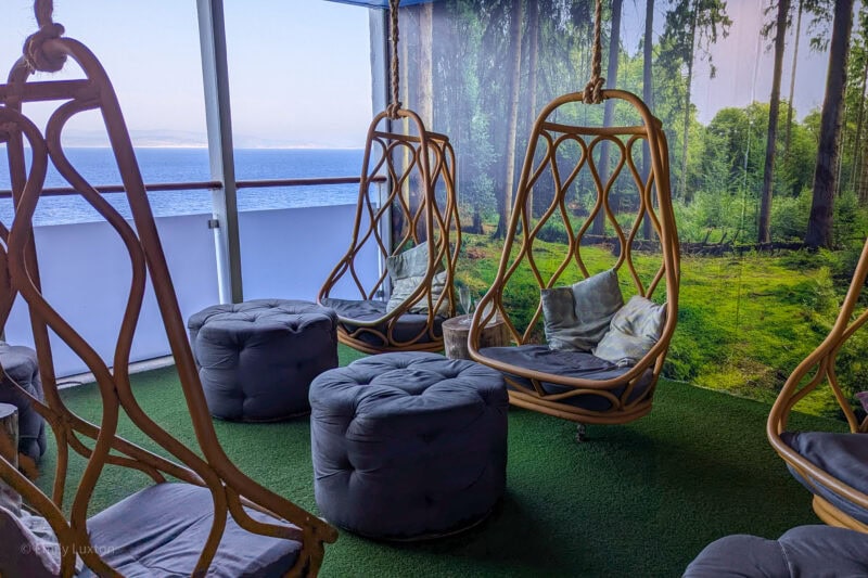 room with fake grass floor and several wooden oval shaped swinging chairs with grey cushions. There are two grey circular pouffes between them and a glass wall with a view of the ocean. 