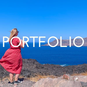 Button. The text reads Portfolio in white letters. The photo behind shows Emily in a long red dress being swept up by the wind standing on a cliff edge with bright blue sea and a grey rocky island beyond.