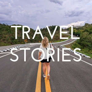 Buttone. The text reads Travel Stories in white. It is laid over a photo of Emily wearing a white shirt and dark blue denim shorts walking away from the camera along a road which winds away into the distance. 