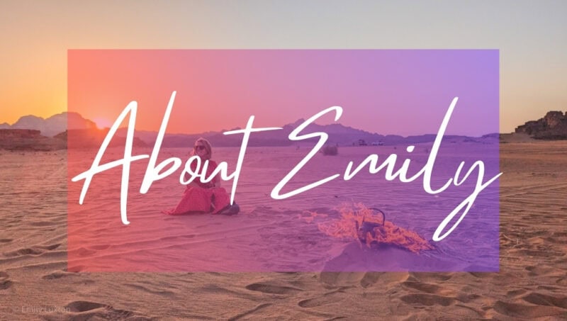 About Emily written in white text over a pink opague box on top of a phot. The photo shows British travel blogger Emily Luxton sitting in the desert next to a fire with the sun setting behind her. She is wearing a long red ress and sunglasses and there is an iron kettle on the campfire.