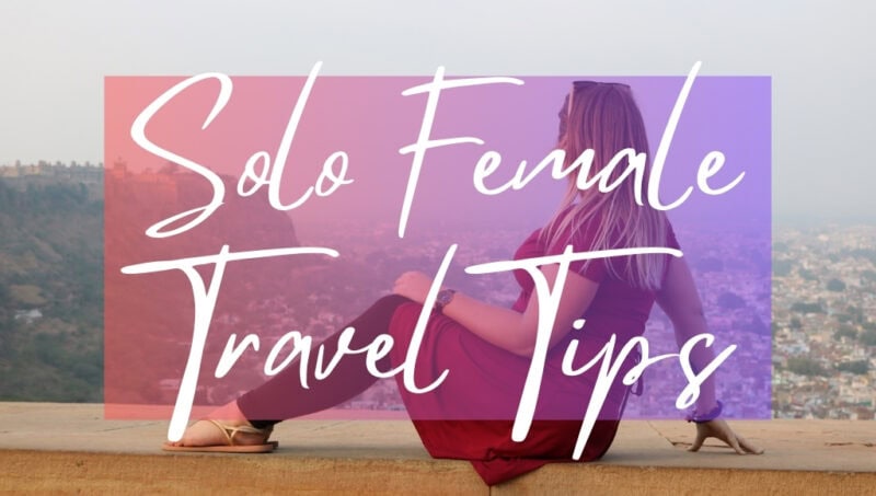 Solo Female Travel Tips in white writing on a pink opague box, on top of a photo showing solo female travel blogger Emily Luxton wearing a dark red dress facing sitting on a wall. She is looking away from the camera and has her long blonde hair down. The view behind is of a city skyline out of focus.