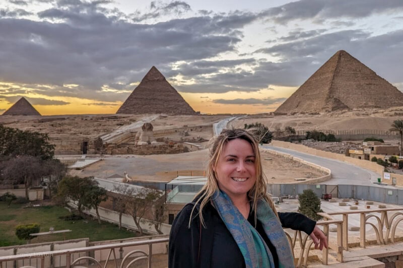 Travel blogger Emily Luxton standing on a balcony with a view of the three pyramids of Giza behind her. It is just after sunset and the sky is cloudyw ith a golden glow on the horizon. Emily is wearing a black cardigan with grey trainers and a green scarf.