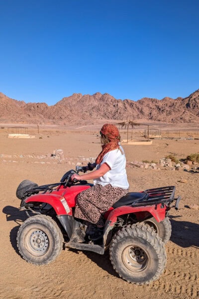 Solo female travel blogger Emily Luxton on a quad bike in the desert in Sharm el Sheikh Egypt. There is a sandy desert landscape behind her with rugged, rocky mountains and a clear, cloudless blue sky. Emily is wearing leopard print wide-leg trousers and a white shirt and has an orange scarf wrapped around her face and hair in the style of a bedouin.