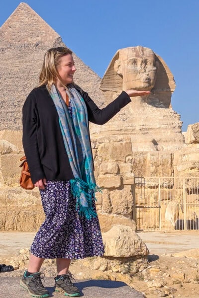 Emily standing on a rock in front of the Sphinx in Egypt, with her hand held out palm flat. The camera angle makes it look like she is petting the Sphinx's chin. Emily is wearing a long blue maxi dress with white flowers and a black cardigan with grey trainers and a green scarf. One of the pyramids is visible behind them.